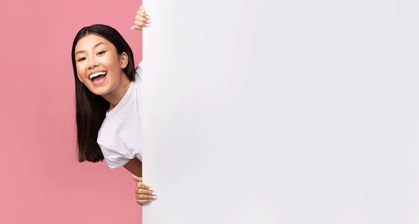 Menina asiática alegre espreitando fora do cartaz branco, fundo rosa — Fotografia de Stock