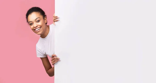 Meisje gluuren uit blanco poster staande over roze achtergrond — Stockfoto