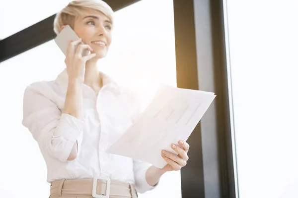 Pani przedsiębiorca rozmawia przez telefon stojąc w pobliżu okna w biurze — Zdjęcie stockowe