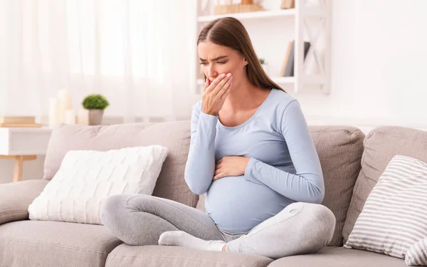 Беременная девушка, касающаяся рта, сидит на диване в помещении — стоковое фото