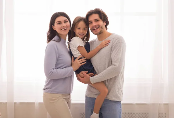 Joyful millennial parents posing against window with little daughter on hands — 스톡 사진