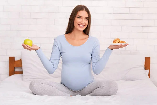 Mulher grávida segurando maçã e Croissant sentado na cama — Fotografia de Stock