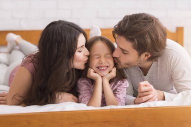 Sevgi dolu anne ve baba yatakta küçük kızlarını öpüyor.