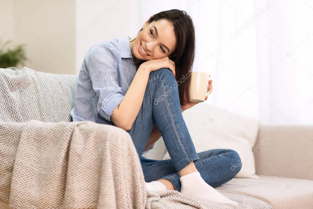 Cheerful young girl sitting on sofa with cup of coffee