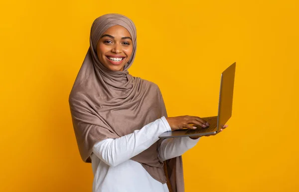 Boldog fekete muszlim nő hidzsáb gazdaság laptop, sárga stúdió háttér — Stock Fotó