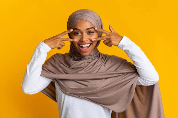 Fröhliches afro-muslimisches Mädchen mit Kopftuch, das V-Zeichen-Symbole in Augennähe zeigt — Stockfoto