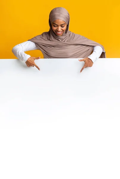 Mujer afro musulmana en pañuelo para la cabeza apuntando al tablero de anuncios blanco — Foto de Stock