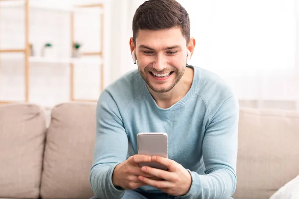 Hombre alegre en los auriculares usando Smatphone sentado en el sofá interior —  Fotos de Stock