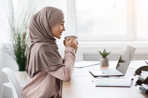 Muszlim üzletasszony kávét iszik a munkahelyen, miközben laptopot használ az irodában — Stock Fotó