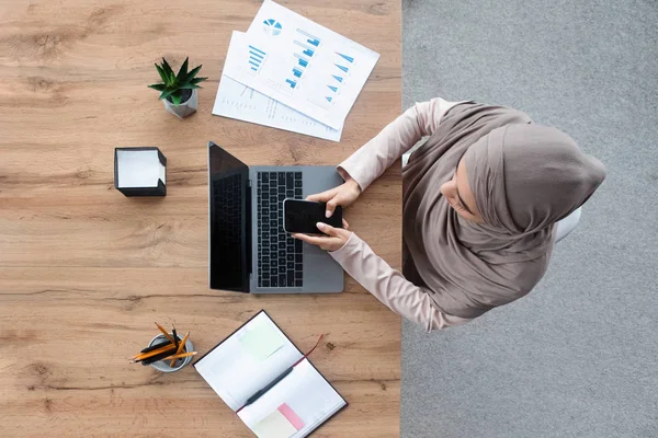 Arabisch vrouwelijke werknemer met behulp van smartphone en werken op laptop, Top View — Stockfoto