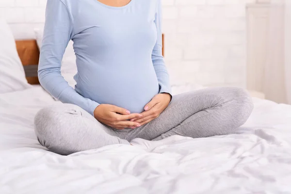 Chica embarazada sentada en la cama tocando su vientre interior, recortado — Foto de Stock