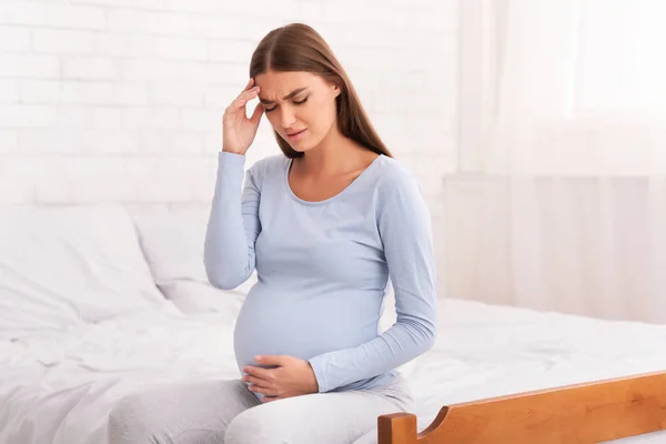 Gravid kvinna som har huvudvärk dåligt sittande på sängen inomhus — Stockfoto