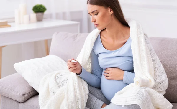 Нещаслива вагітна леді вимірювання температури сидячи на дивані вдома — стокове фото
