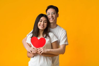 Asyalı çift sarılıyor ve kocaman kırmızı bir kalp taşıyor.