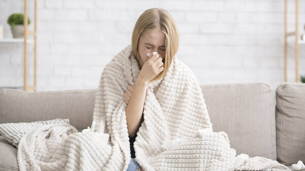 Молодая девушка заболела, завернутая в одеяло, сморкалась в нос
