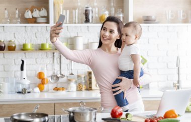Mutlu bir anne ve bebek mutfakta cep telefonuyla selfie çekiyor.