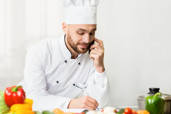 Maschio chef parlare su cellulare prendere appunti in piedi in cucina — Foto Stock