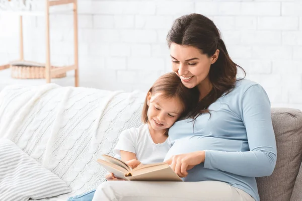 Underbar liten flicka läser bok med sin gravida mamma — Stockfoto