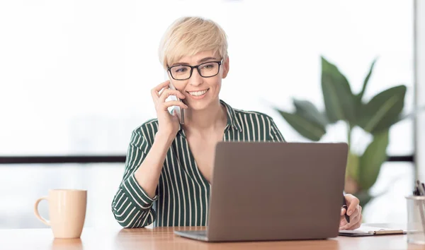 Женщина разговаривает по мобильному телефону с помощью ноутбука, работающего в офисе — стоковое фото