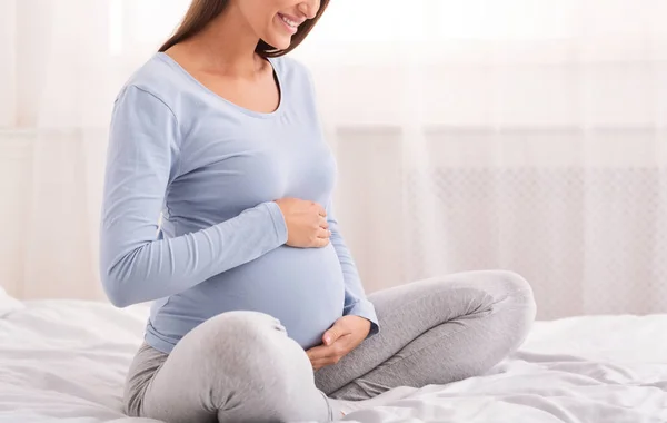 Terhes lány mosolygós ül az ágyon a kényelmes hálószoba, vágott — Stock Fotó