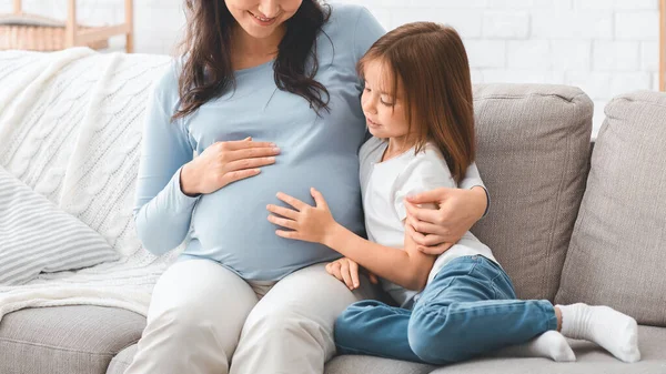 Girl sitting on sofa with pregnant mom, touching her belly — 스톡 사진