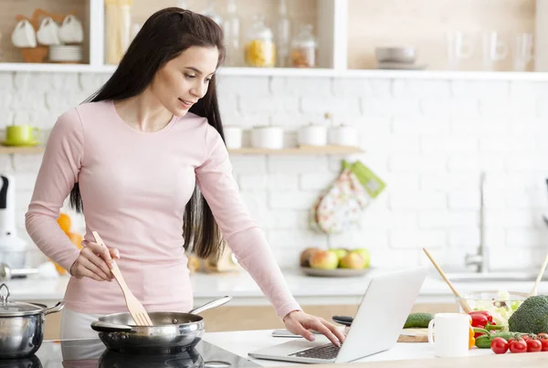 Millennial vrouw in keuken koken met verwijzing naar de laptop — Stockfoto