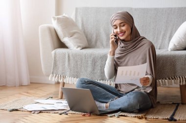Kadın serbest çalışan, kağıtları kontrol eden, cep telefonuyla konuşan ve dizüstü bilgisayarla çalışan.