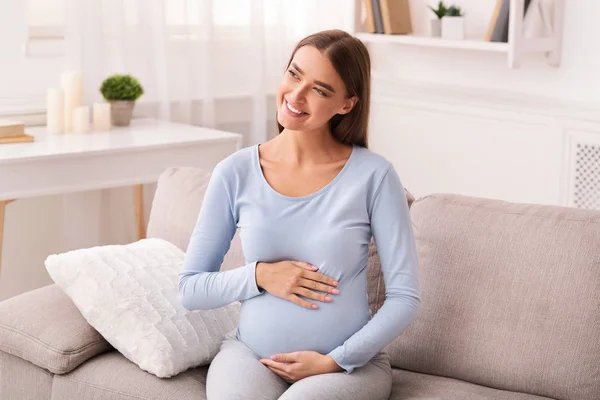 Vidám terhes hölgy megérinti has ül a kanapén otthon — Stock Fotó