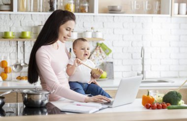 Genç anne dizüstü bilgisayarda yazıyor, bebekle mutfakta oturuyor.