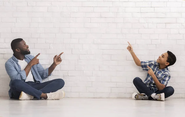 Svart far och son sitter på golvet nära vit tegelvägg och pekar på tom rymd — Stockfoto