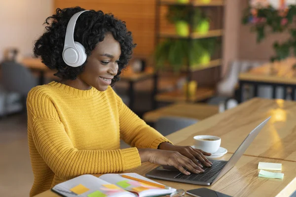 Chica negra feliz en los auriculares mirando a la computadora portátil, café interior —  Fotos de Stock