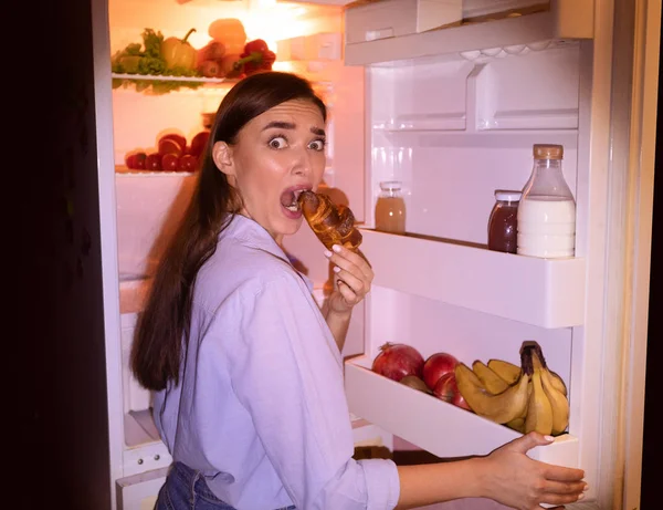 Vystrašená dívka v pyžamu jíst croissant v blízkosti otevřené ledničky — Stock fotografie