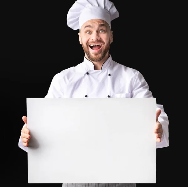 Emocionado chico cocinero sosteniendo tablero blanco de pie sobre fondo negro — Foto de Stock