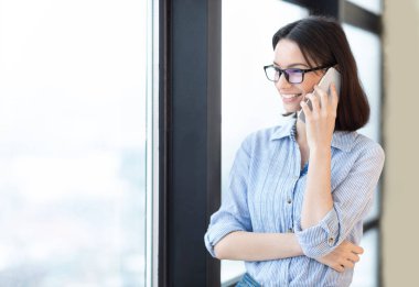 Gülümseyen kız cep telefonuyla pencerenin yanında konuşuyor.