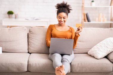Dizüstü bilgisayarı ve kredi kartıyla koltukta oturan Afro Kadın