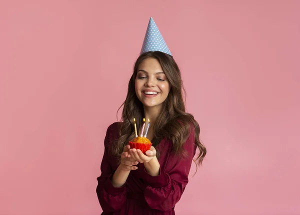 Glädjande ung kvinna i födelsedagshatt håller cupcake med tända ljus — Stockfoto