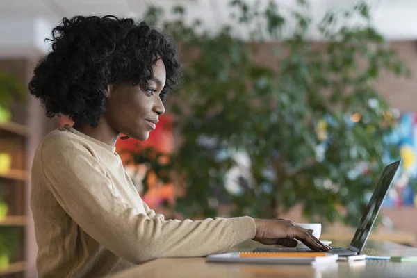 Kafede çalışan genç afro kadın, dizüstü bilgisayar kullanıyor. — Stok fotoğraf