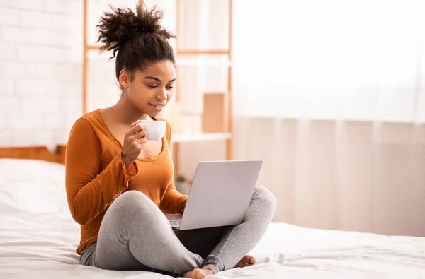 Afroamericano signora utilizzando computer portatile avendo caffè seduto su letto — Foto Stock