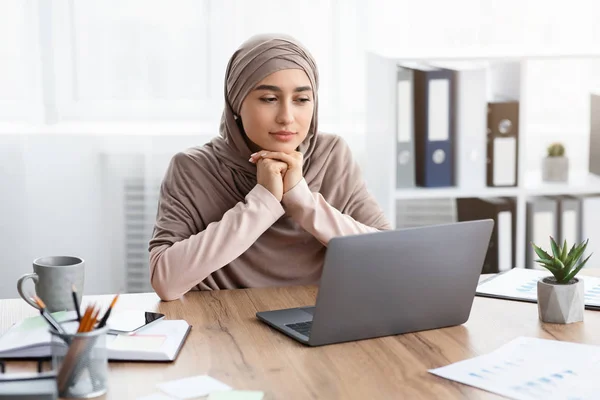 Задумчивая мусульманка-бизнесмен сидит на рабочем месте и смотрит на экран ноутбука — стоковое фото