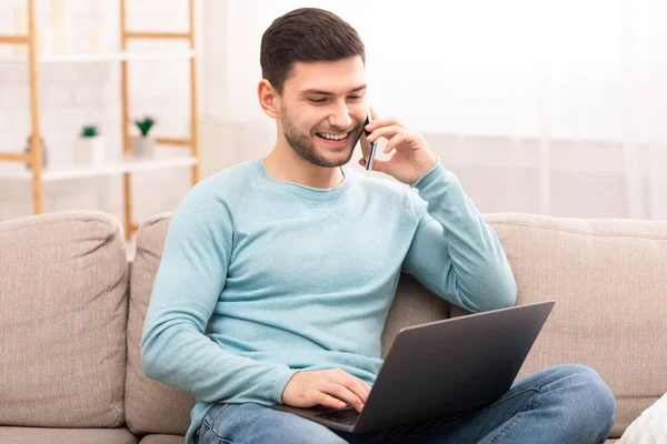 Tipo hablando por teléfono trabajando en el ordenador portátil sentado en el sofá — Foto de Stock