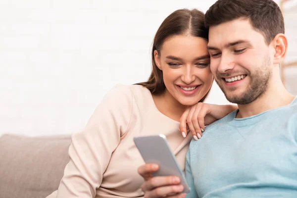 Casal sorridente usando telefone celular sentado no sofá interior — Fotografia de Stock