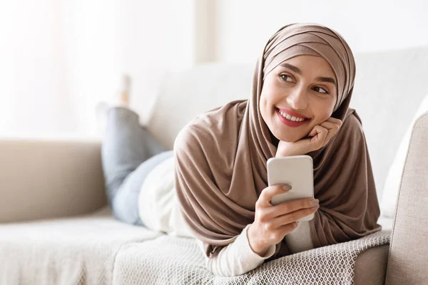 Gadis arab romantis dalam jilbab santai di sofa dengan smartphone — Stok Foto