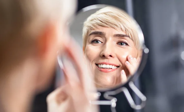 Женщина средних лет, глядя в зеркало, касается гладких щек в ванной комнате — стоковое фото