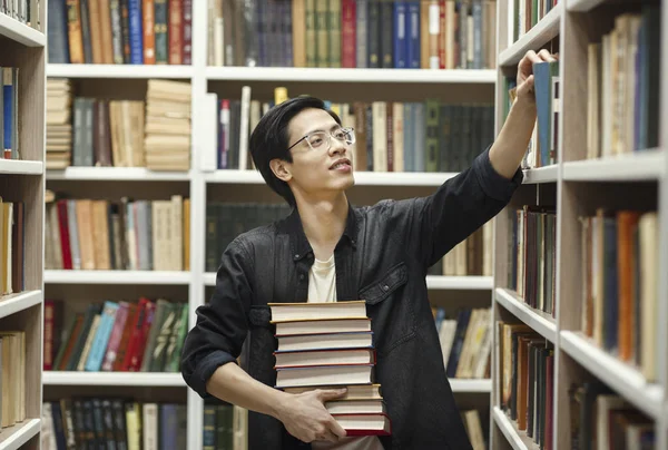 Young chinese man in glasses picking book at library — Stockfoto