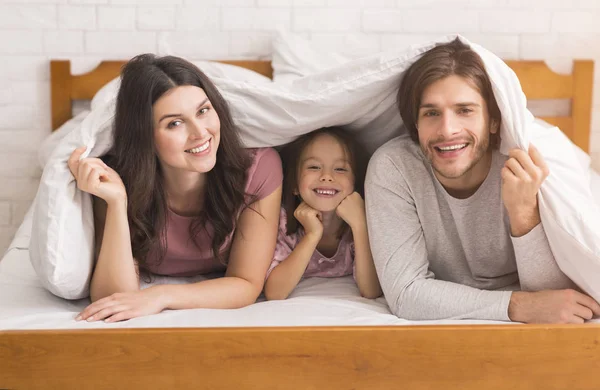 Mamma, papà e carino piccolo figlia nascondendo sotto coperta su letto — Foto Stock