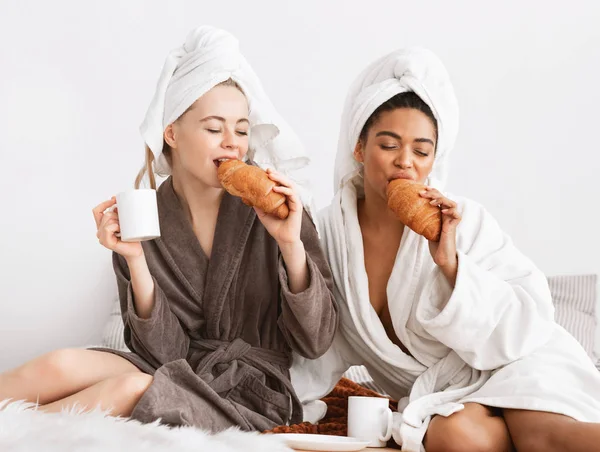 Multiethnic spa girlfriends enjoying breakfast, sitting on bed