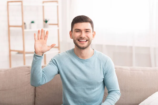 Millennial Guy Waving Hello Smiling To Camera Sitting On Sofa