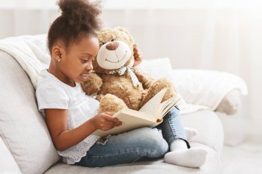 Little afro girl holding teddy bear and reading book
