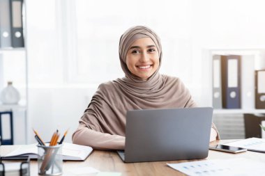 Portrait of happy arabic businesswoman in hijab using laptop at workplace