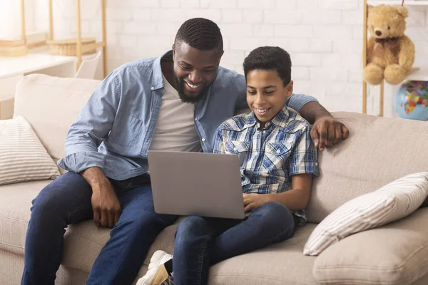 Отец учит сына пользоваться ноутбуком, сидя дома на диване — стоковое фото